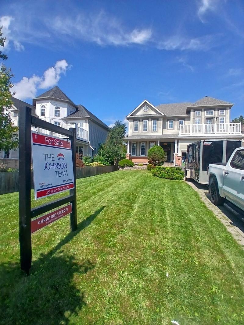 A black double post real estate sign on a Whitby front lawn, with a for sale header strip, a team panel and a name rider along the bottom rail.