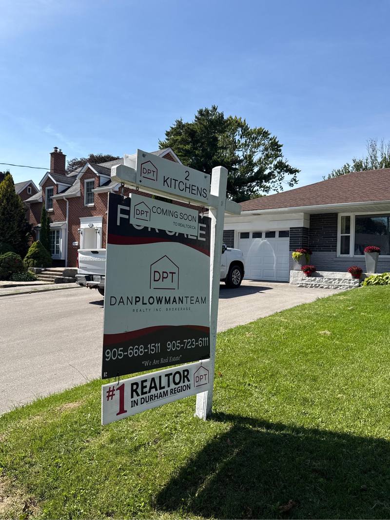 A real estate lawn sign on a white post frame on a front lawn, carrying a top rider, a smaller coming soon panel over the for sale header, and a bottom rider.